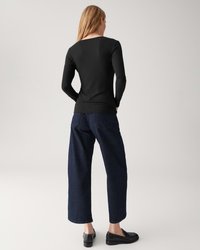 Frau in einem schwarzen Langarmshirt, dunklen weit geschnittenen Jeans und schwarzen Loafers, steht mit den Händen in den Taschen, mit dem Rücken zum Betrachter.