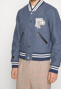 Blå varsityjacka i ullblandning med randig ribbad krage och muddar, med en framsides-logopatch och svarta fusk läder sidofickor.