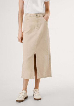Kvinde iført en beige midi-nederdel med frontslids og flettet bælte, parret med hvide sneakers og en hvid top med korte ærmer.