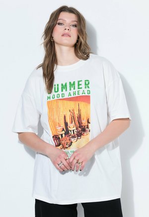 Joven con el pelo de longitud media lleva una camiseta blanca de gran tamaño con el texto "SUMMER MOOD AHEAD" y un estampado de cactus, posando frente a una pared blanca.