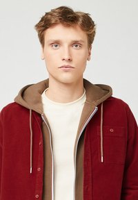 Giacca camicia in velluto rosso con una felpa con cappuccio marrone sotto; presenta un collo a giro bianco e una tasca frontale. Tessuto dalla texture liscia.