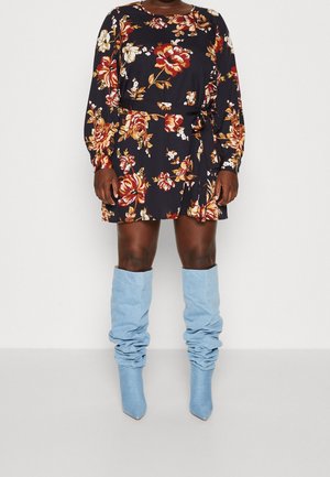 Abito mini floreale con maniche lunghe, sfondo blu navy, fiori rossi e gialli, vita cintrata con cintura. Stivali slouch fino al ginocchio di colore azzurro chiaro.