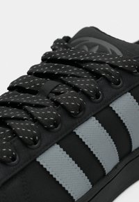 Μαύρα αθλητικά παπούτσια με υφή συνθετικού υλικού, διακοσμημένα με γκρι ρίγες, επίπεδα κορδόνια με αντανακλαστικές κλωστές και αντίθετες λεπτομέρειες.