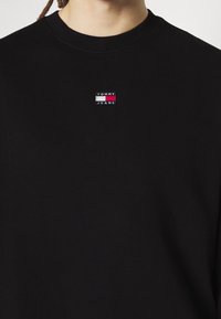 Sweat-shirt noir à col rond, avec un petit logo rectangulaire Tommy Jeans en rouge, blanc et bleu cousu sur la poitrine.