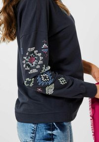 Felpa blu navy con motivi decorativi in paillettes nei toni del blu, verde e rosso sulla manica sinistra. Polsini e orlo a coste, tessuto morbido.