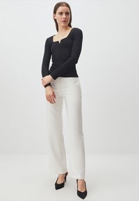 Maglietta nera a maniche lunghe con scollatura quadrata e dettagli arricciati, abbinata a pantaloni bianchi a vita alta e scarpe con tacco nere.
