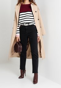 Beżowy trencz na bordowo-białym swetrze w paski, czarne dżinsy i bordowe botki za kostkę; trzyma okrągłą, ciemną torebkę.