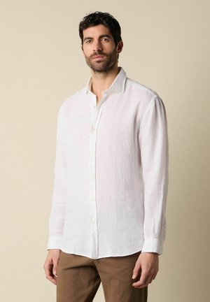 Mann mit kurzen dunklen Haaren und Bart, trägt ein weißes Hemd mit langen Ärmeln und braune Hose, steht vor einem beigen Hintergrund.