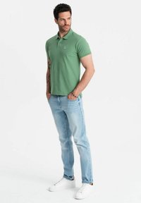 Polo vert à manches courtes, jean bleu clair et baskets blanches. Le polo présente un logo discret sur la poitrine.