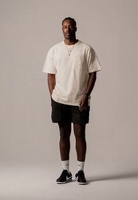 Krém színű pamut póló, fekete rövidnadrág, fekete sportcipők és fehér bokazokni. Egyszerű dizájn zsebdísszel a pólón.