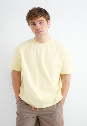 Giovane uomo con capelli ricci che indossa una maglietta giallo pallido e pantaloni marroni, in piedi con le mani nelle tasche davanti a uno sfondo bianco.