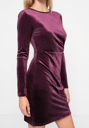 Femme portant une robe en velours violet foncé, ajustée et à manches longues, avec un col haut, debout devant un fond clair uni.