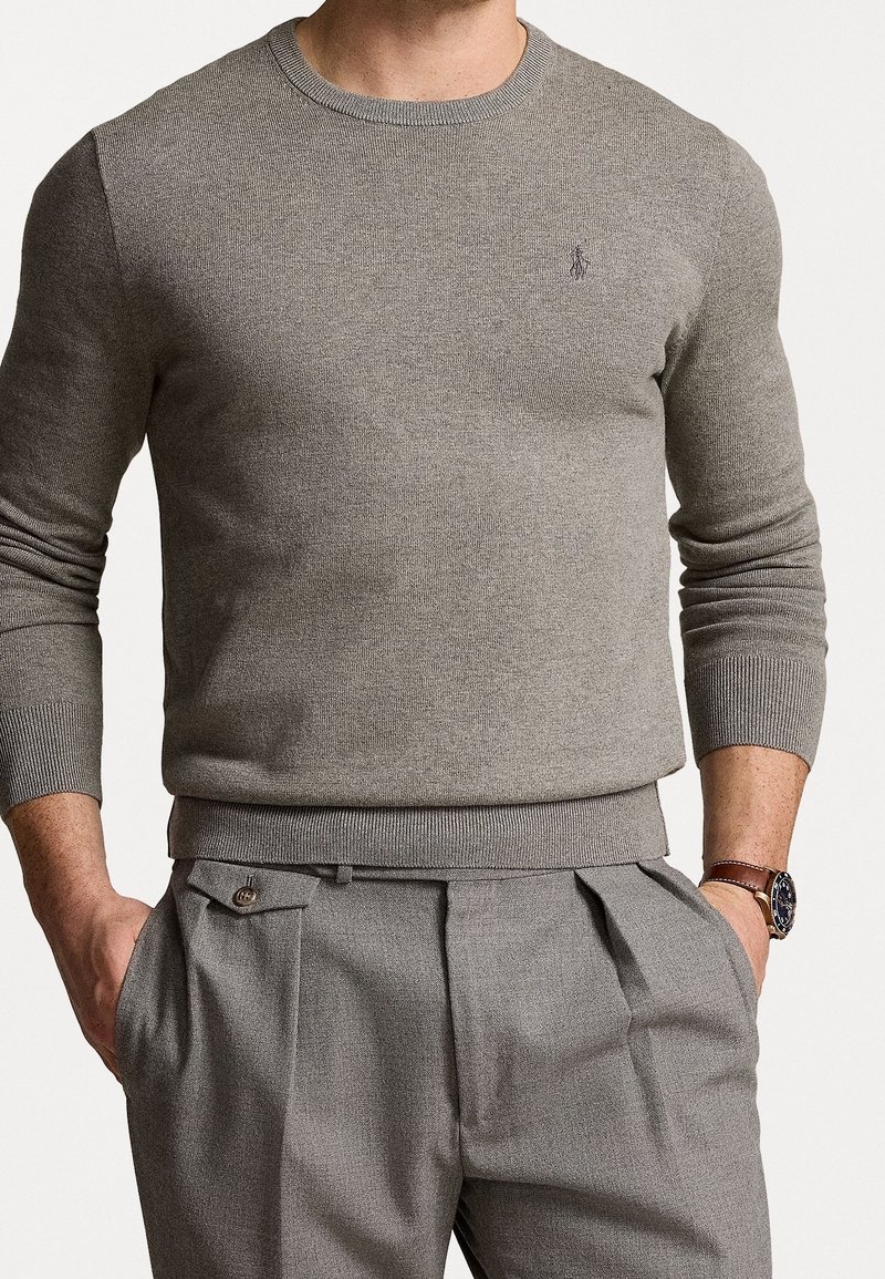 Homme portant un pull col rond ajusté marron clair et un pantalon plissé assorti, les mains dans les poches, montre à bracelet en cuir marron visible.