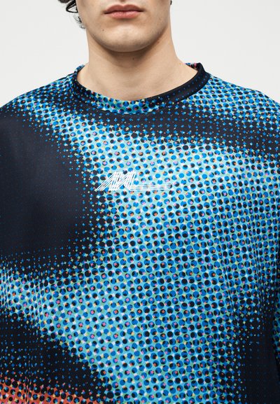 Homme portant une chemise à motifs à pois bleus, noirs et orange avec un petit logo abstrait blanc sur la poitrine, col visible.