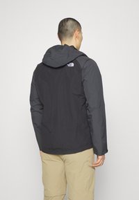 Giacca con cappuccio nera e grigia con finitura resistente all'acqua, dotata di un logo sul retro e polsini regolabili. Tessuto leggero, design ergonomico.