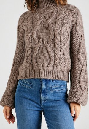 Femme portant un pull épais en maille torsadée couleur taupe avec un col montant et des manches larges, associé à un jean bleu.