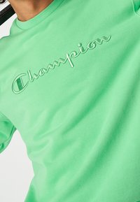 Green shirt with raised embroidered "Champion" logo across the chest, worn by a person with curly dark hair.