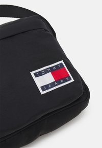 Czarna torba z wypustkami, z białą łatą z niebieskim, czerwonym i białym prostokątnym logo z napisem "TOMMY JEANS" oraz kontrastującymi czarnymi akcentami.