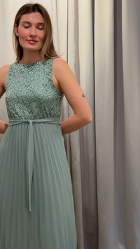 Γυναίκα με μαλλιά μέχρι τους ώμους, που φοράει φόρεμα σε σάπιας πράσινης απόχρωσης χωρίς μανίκια με δαντελωτό μπούστο και πλισέ φούστα, στέκεται μπροστά από μπεζ κουρτίνες.