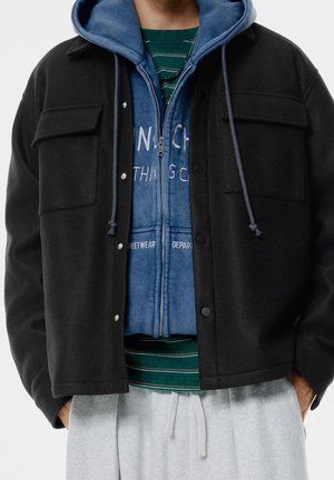 Man draagt een zwarte jas met knopen over een blauwe zip-up hoodie en een groen gestreept overhemd, gecombineerd met lichtgrijze broek met trekkoord.