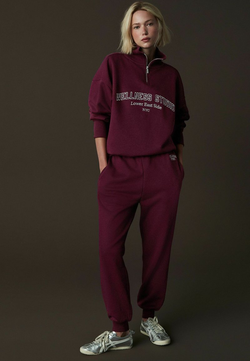 Burgundowa bluza z wysokim kołnierzem i zamkiem, z napisem "WELLNESS STUDIO". Połączona z dopasowanymi burgundowymi dresami i srebrnymi sneakersami.