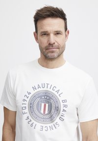 Balta medvilninė marškinėlių su apvaliu logotipu, kuriame užrašyta "Nautical Brand Since 1924", tamsiai mėlynos ir raudonos spalvos akcentais bei trumpomis rankovėmis.