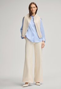 Camicia azzurro chiaro con bottoni, pantaloni cream a gamba larga e un gilet di maglia beige appoggiato sulle spalle, completato da tacchi bianchi appuntiti.