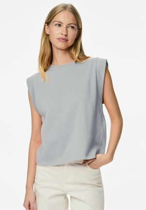 Kobieta o długich blond włosach, ubrana w jasnoszary bezrękawnik i białe spodnie, stoi z jedną ręką w kieszeni, z lekkim spojrzeniem w lewo.