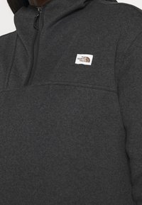 Pullover in pile grigio scuro con colletto a mezza zip, con un piccolo logo bianco sul lato sinistro del petto e una superficie a motivo testurizzato.