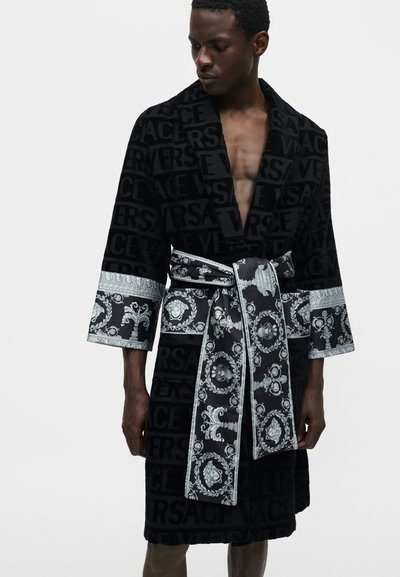 Άνδρας που φοράει μαύρη βελούδινη ρόμπα με ανάγλυφο κείμενο "Versace", φαρδιές διακοσμημένες μανσέτες και ταιριαστή ζώνη με λευκά μπαρόκ σχέδια.