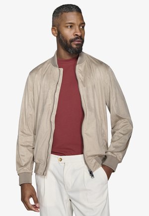 Homme aux cheveux courts et bouclés avec une barbe, portant une veste aviateur beige sur une chemise rouge et un pantalon blanc, debout avec une main dans la poche.