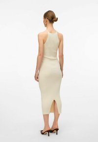Ribbet, beige midi-kjole med tynde skulderstropper og en slids i ryggen. Glat tekstur med subtile lodrette striber, parret med hælede sandaler.