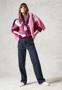 Randig stickad tröja i vinröd, krämfärgad och rosa; löst sittande mörk denimjeans; vita sneakers. Halsduk hängande över axlarna. Studiebakgrund.