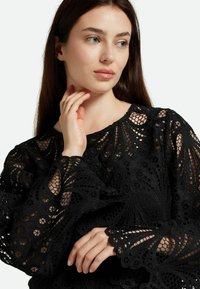Blusa nera in pizzo con intricate aperture, maniche lunghe a campana e scollatura tonda. Tessuto morbido con dettagli di design elaborati.