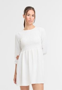 Witte jurk met lange mouwen, met een gesmockte bodice met textuur en een vloeiende rok. Lichtgewicht stof met een subtiele glans.