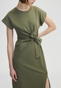 Olivengrünes Kleid aus weichem Stoff, mit Rundhalsausschnitt, kurzen Ärmeln und einem Knoten-Detail an der Taille sowie einem seitlichen Schlitz.