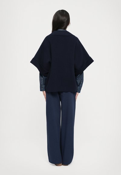 Marineblauwe wollen poncho in een mixstof met brede mouwen, laag over een donkerdenim shirt. Losse snit, met minimale details voor een strakke silhout. 