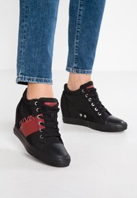 Fötter i svarta kilklackssneakers med röd rand och "Calvin"-text, i kombination med blå denimjeans, som står på vit golv.