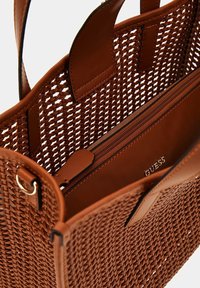Sac fourre-tout tissé marron avec des accents en cuir lisse, doté d'une poche zippée à l'intérieur et du nom de la marque "GUESS" estampillé sur la doublure.