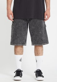 Shorts cargo en denim noir avec poches latérales, ourlets effilochés et délavage, associés à des baskets noires et des chaussettes blanches avec logo.