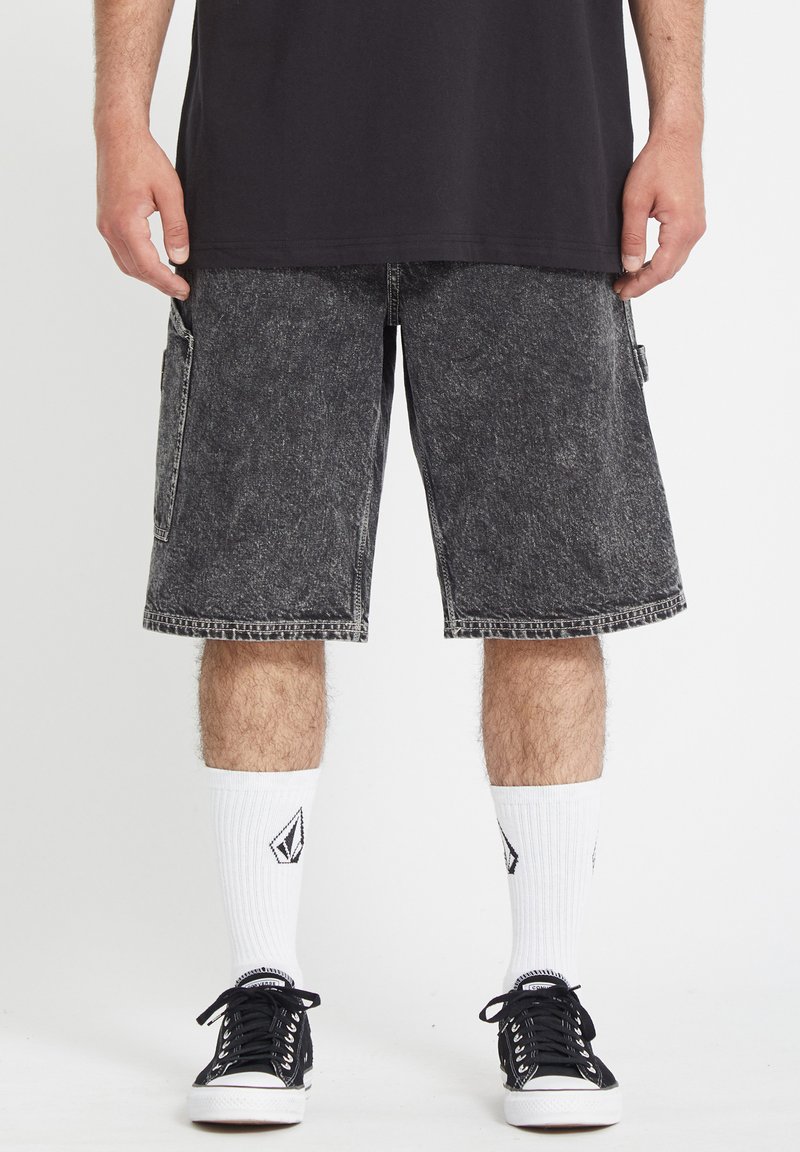 Shorts cargo en denim noir avec poches latérales, ourlets effilochés et délavage, associés à des baskets noires et des chaussettes blanches avec logo.