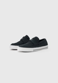 Sorte semskede slip-on sko med rund tå, hvit gummisåle og detaljer, med en frontstropp og teksturert overflate.