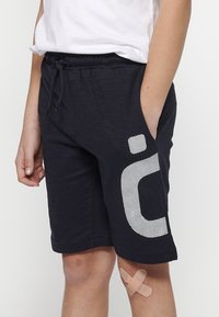 Marineblaue Shorts aus weichem Stoff, mit einem großen weißen gestreiften Logo am linken Bein und zwei seitlichen Taschen. Ein Verband ist am Knie sichtbar.