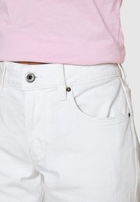 Shorts in denim bianco con un taglio classico, dotati di chiusura frontale con bottone, tasche laterali e dettagli di cucitura discreti. Abbinati a una maglietta rosa.