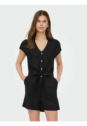 Jeune femme portant une combinaison noire à manches courtes avec boutons et ceinture nouée à la taille, debout les mains dans les poches sur un fond uni.