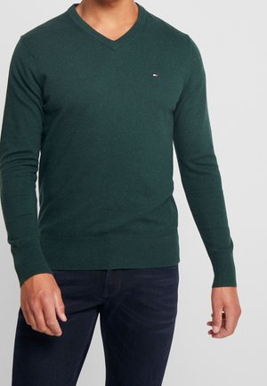 Mand iført en mørkegrøn V-hals sweater med lille logo på brystet og mørkeblå jeans, stående mod en ensfarvet lys baggrund.