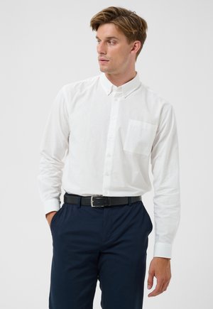 Hombre con camisa blanca abotonada con un bolsillo en el pecho, metida dentro de pantalones azul oscuro con cinturón negro, mirando hacia su izquierda.