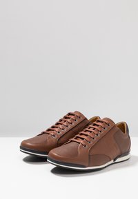 Baskets en cuir marron avec une finition texturée, des accents en mesh, une semelle blanche et des détails gris contrastants sur le talon et les côtés.