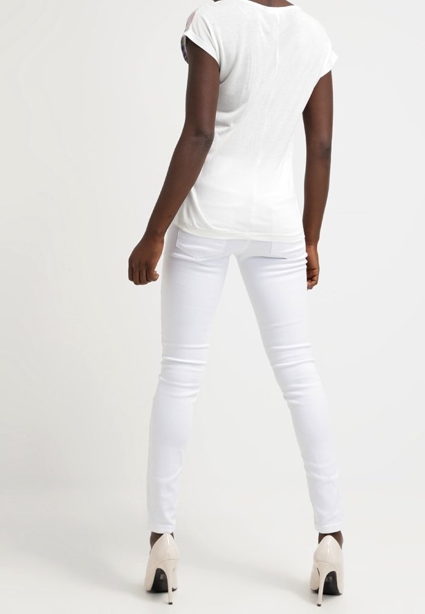 Femme debout de dos face au spectateur portant un haut blanc à manches courtes, un pantalon skinny blanc et des chaussures à talons beiges sur un fond uni.