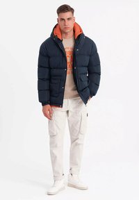 Chaqueta tipo plumón en navy con forro naranja, que cuenta con capucha, cierre de cremallera y bolsillos frontales. Llevada sobre una sudadera gráfica beige y pantalones cargo blancos.